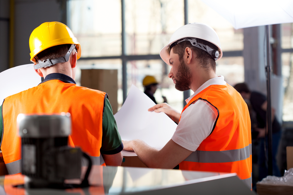 Two civil engineers wearing hard hats and high-visibility vests check construction plans in an industrial or construction site environment. The image stands for project management in construction, project control and cooperation in infrastructure projects.