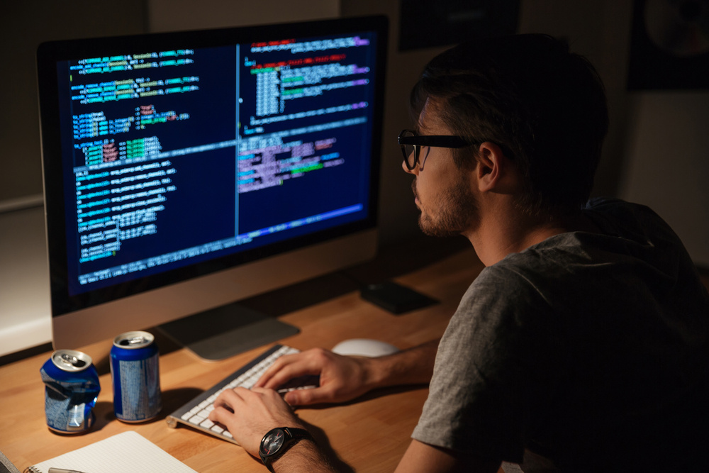 A young man wearing glasses works intently at a desk in front of a large computer screen displaying multiple windows with colored programming code. Two beverage cans and a notepad are on the desk.