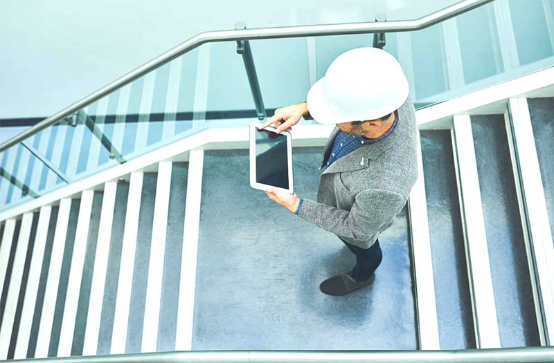 A construction manager wearing a white safety helmet stands on a staircase in a modern building, using a tablet for digital project monitoring.