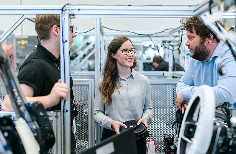 Drei Ingenieure und Fachkräfte diskutieren in einer Produktionsumgebung mit Maschinen und technischen Anlagen. Das Bild zeigt Zusammenarbeit im industriellen Umfeld sowie Themen wie Digitalisierung, Industrie 4.0 und Projektmanagement.