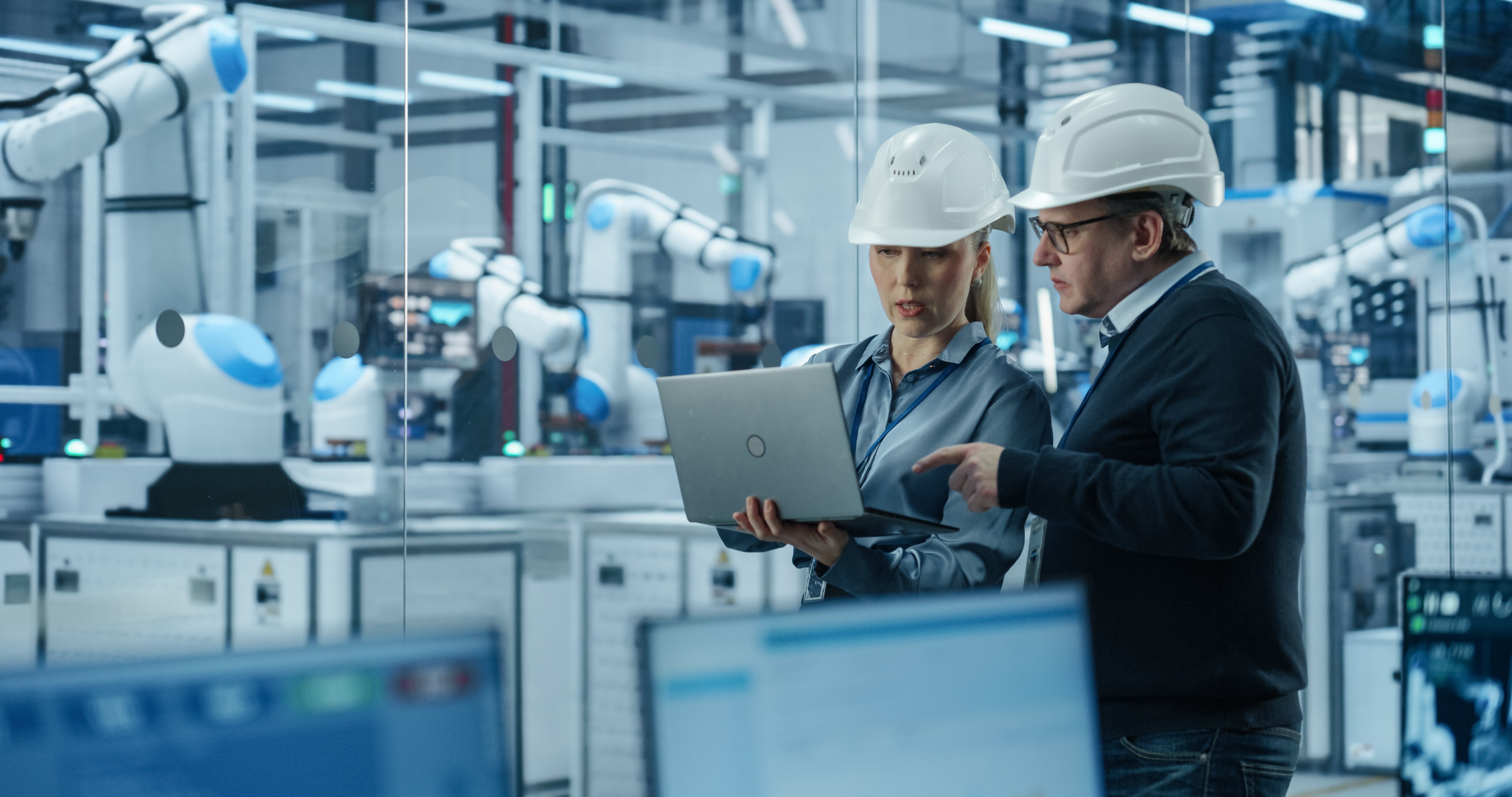 Zwei Ingenieure mit Schutzhelmen analysieren Produktionsdaten auf einem Laptop in einer modernen Industrie-4.0-Fabrik mit automatisierten Robotersystemen. Im Hintergrund sind vernetzte Fertigungsanlagen und kollaborative Roboterarme zu sehen. Das Bild steht für digitale Transformation, Smart Factory, industrielle Automatisierung sowie datengetriebene Projektsteuerung und effiziente Produktionsprozesse in der Fertigungsindustrie.