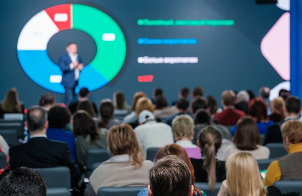 Konferenzpublikum von hinten, das einem Redner zusieht, der ein Diagramm präsentiert